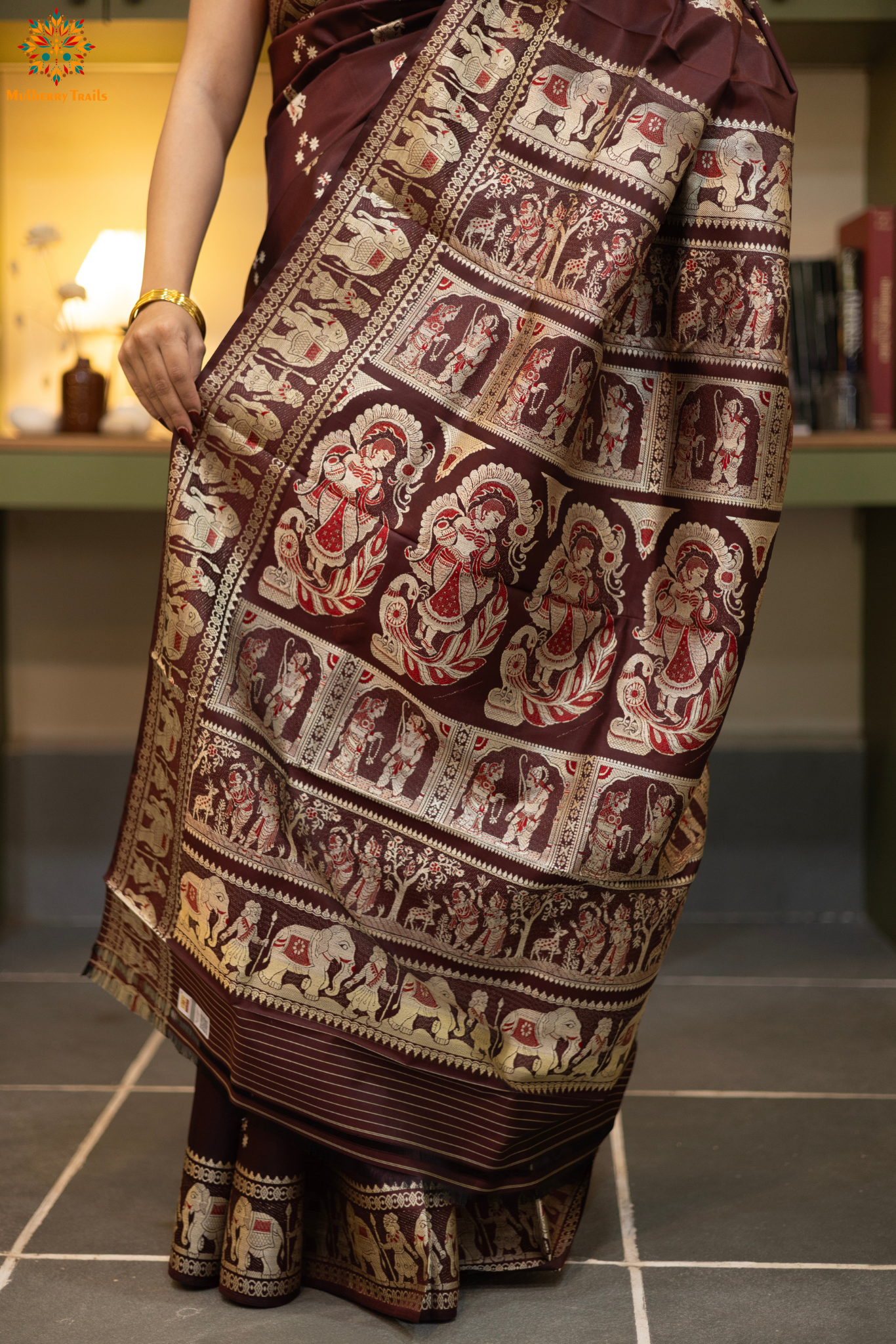Woman in a Coffee brown and silver resham work saree standing indoors with a desk and lamp in the background.
Woman wearing a Coffee brown saree with a pattern in a elegant setting.
Baluchari – Pure Silk Meenakari Saree
1. “Pure silk Baluchari Meenakari saree with meenakari motif weave and Silver resham detailing for a regal look.”
2. “Silk Mark pure silk meenakari saree featuring intricate gold resham work motifs and rich texture.”