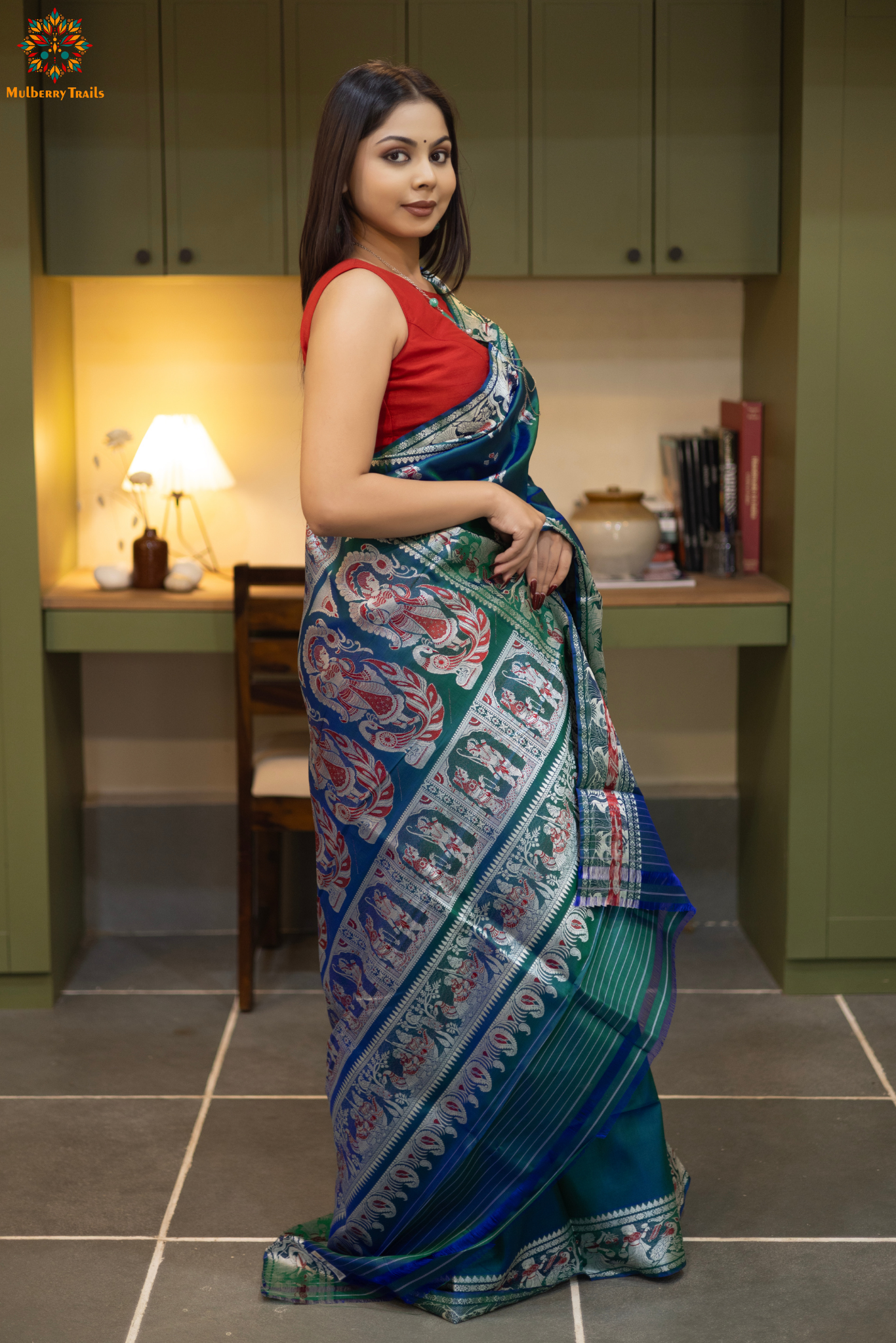 Woman in a Peacock blue and silver resham work saree standing indoors with a desk and lamp in the background.
Woman wearing a Peacock blue saree with a pattern in a elegant setting.
Baluchari – Pure Silk Meenakari Saree
1. “Pure silk Baluchari Meenakari saree with meenakari motif weave and Silver resham detailing for a regal look.”
2. “Silk Mark pure silk meenakari saree featuring intricate gold resham work motifs and rich texture.”