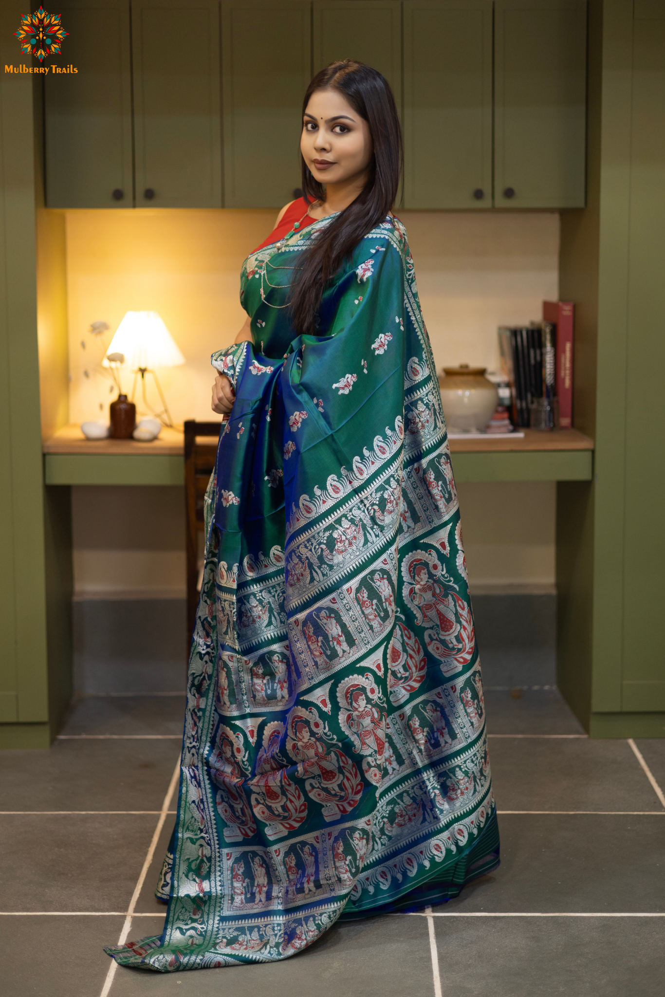 Woman in a Peacock blue and silver resham work saree standing indoors with a desk and lamp in the background.
Woman wearing a Peacock blue saree with a pattern in a elegant setting.
Baluchari – Pure Silk Meenakari Saree
1. “Pure silk Baluchari Meenakari saree with meenakari motif weave and Silver resham detailing for a regal look.”
2. “Silk Mark pure silk meenakari saree featuring intricate gold resham work motifs and rich texture.”