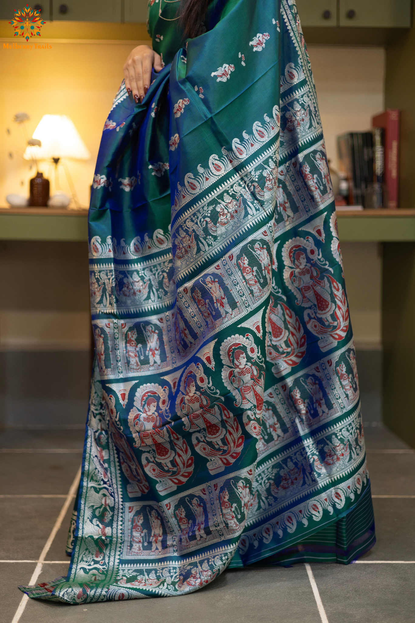 Woman in a Peacock blue and silver resham work saree standing indoors with a desk and lamp in the background.
Woman wearing a Peacock blue saree with a pattern in a elegant setting.
Baluchari – Pure Silk Meenakari Saree
1. “Pure silk Baluchari Meenakari saree with meenakari motif weave and Silver resham detailing for a regal look.”
2. “Silk Mark pure silk meenakari saree featuring intricate gold resham work motifs and rich texture.”