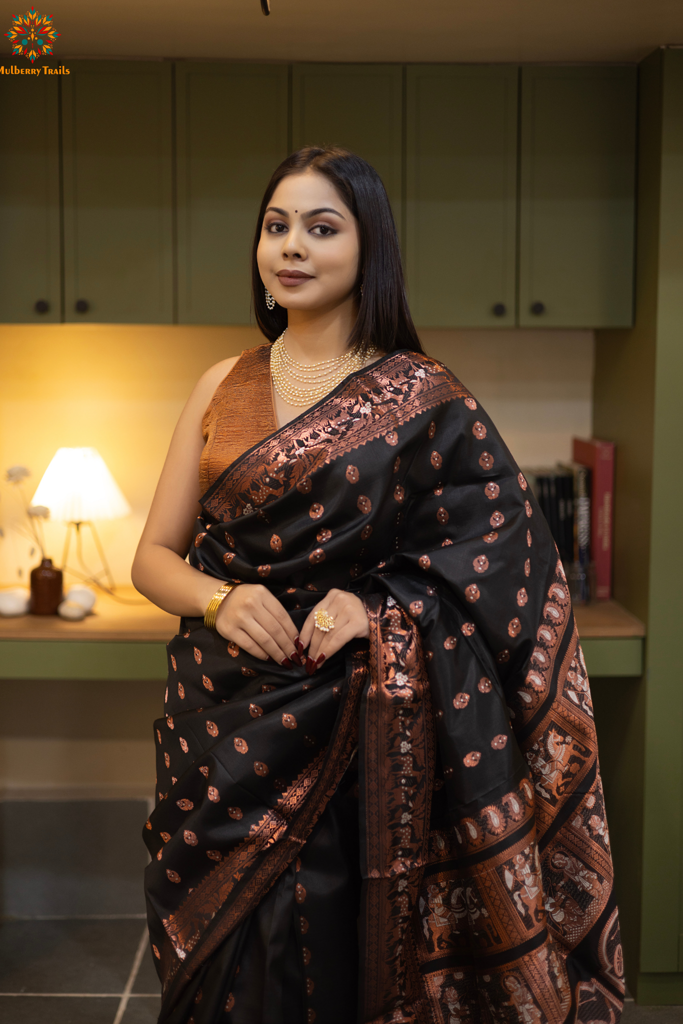 Swarnachari – Pure Silk Hazarbutta Saree
1. “Pure silk Swarnachari saree with hazarbutta thousand-motif weave and copper zari detailing for a regal look.”
2. “Silk Mark pure silk hazarbutta saree featuring intricate copper zari motifs and rich texture.”
Woman wearing a black and copper zari saree in a room with a desk and lamp.