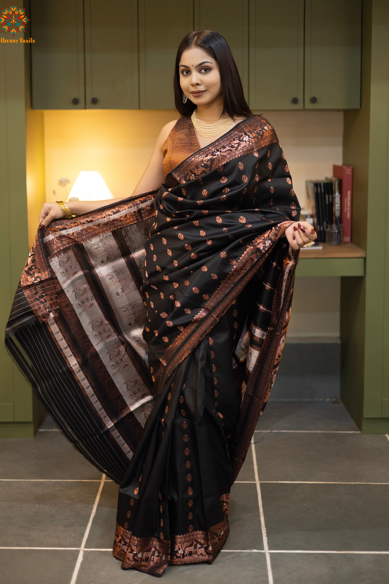 Swarnachari – Pure Silk Hazarbutta Saree
1. “Pure silk Swarnachari saree with hazarbutta thousand-motif weave and copper zari detailing for a regal look.”
2. “Silk Mark pure silk hazarbutta saree featuring intricate copper zari motifs and rich texture.”
Woman wearing a black and copper zari saree in a room with a desk and lamp.