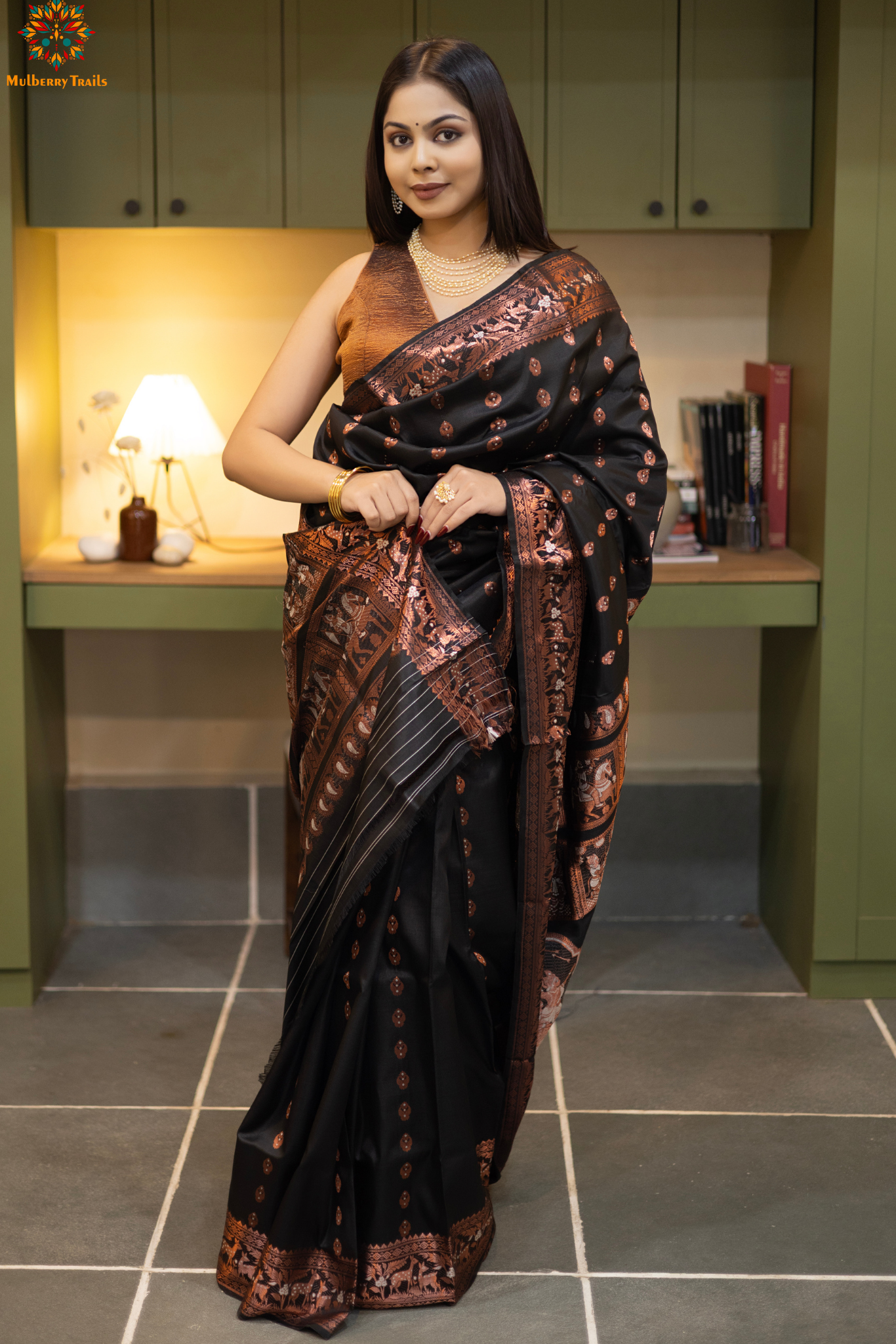 Swarnachari – Pure Silk Hazarbutta Saree
1. “Pure silk Swarnachari saree with hazarbutta thousand-motif weave and copper zari detailing for a regal look.”
2. “Silk Mark pure silk hazarbutta saree featuring intricate copper zari motifs and rich texture.”
Woman wearing a black and copper zari saree in a room with a desk and lamp.
