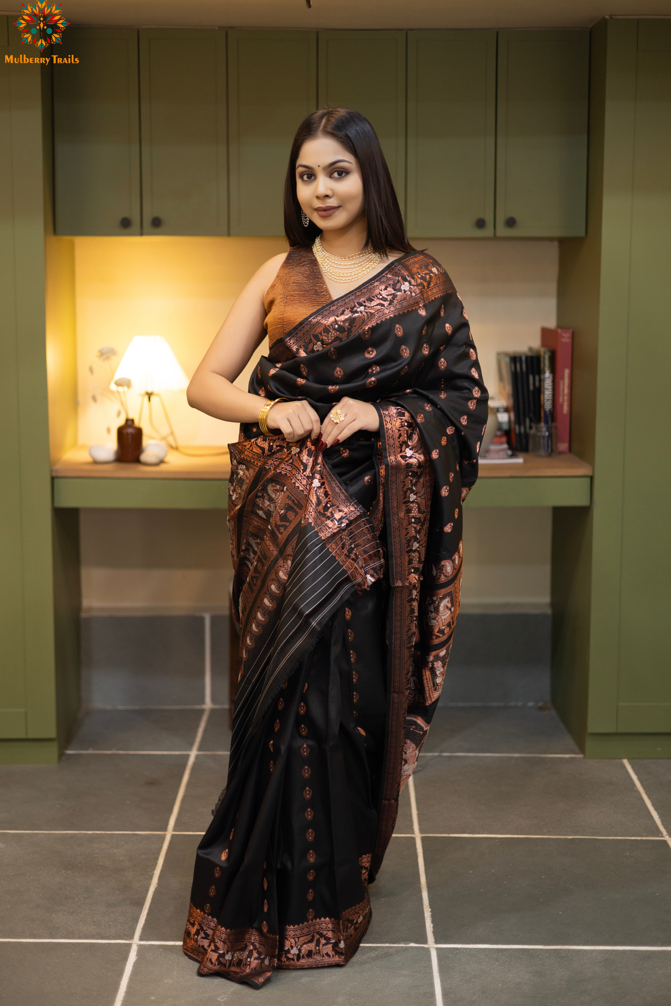 Swarnachari – Pure Silk Hazarbutta Saree
1. “Pure silk Swarnachari saree with hazarbutta thousand-motif weave and copper zari detailing for a regal look.”
2. “Silk Mark pure silk hazarbutta saree featuring intricate copper zari motifs and rich texture.”
Woman wearing a black and copper zari saree in a room with a desk and lamp.