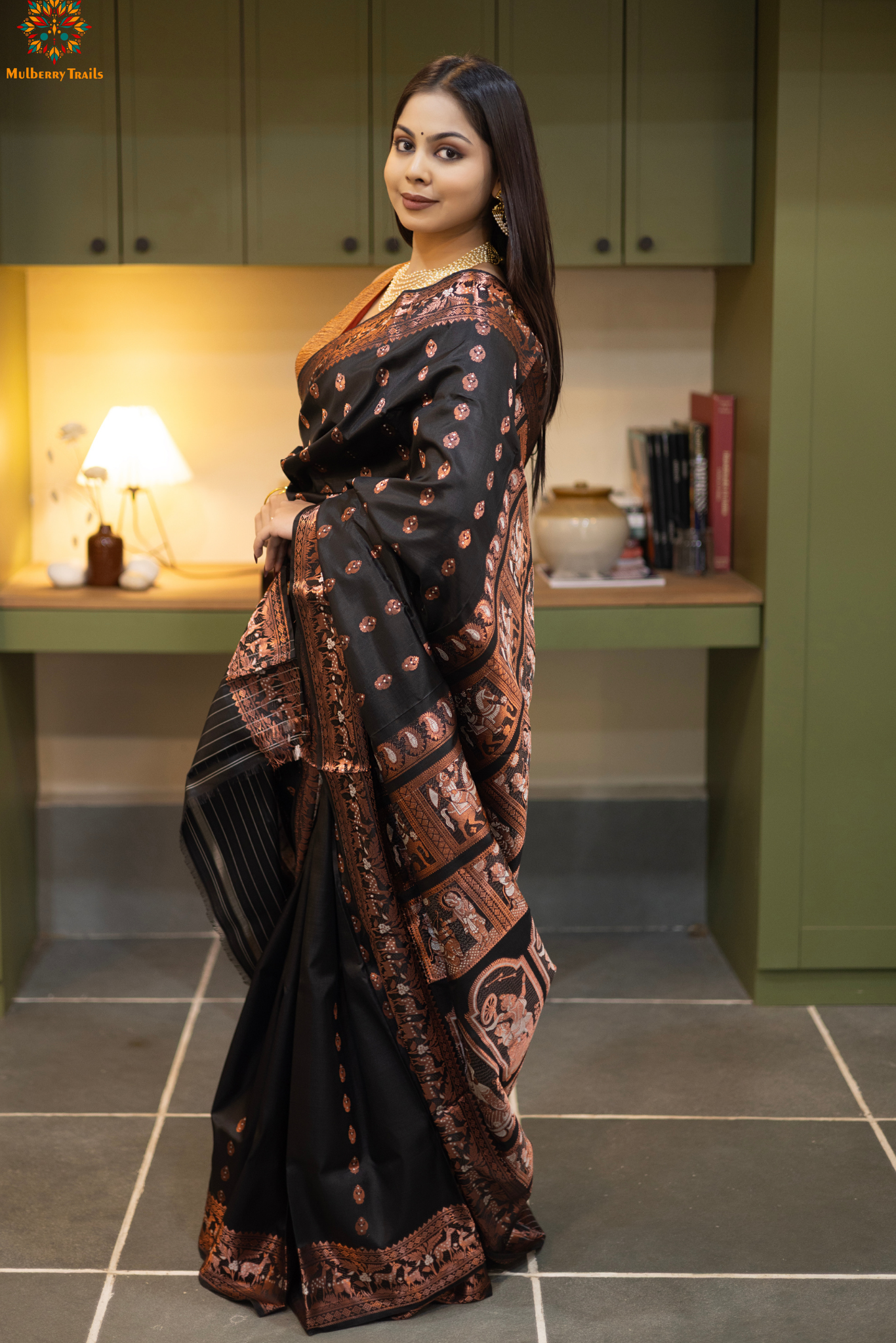 Swarnachari – Pure Silk Hazarbutta Saree
1. “Pure silk Swarnachari saree with hazarbutta thousand-motif weave and copper zari detailing for a regal look.”
2. “Silk Mark pure silk hazarbutta saree featuring intricate copper zari motifs and rich texture.”
Woman wearing a black and copper zari saree in a room with a desk and lamp.