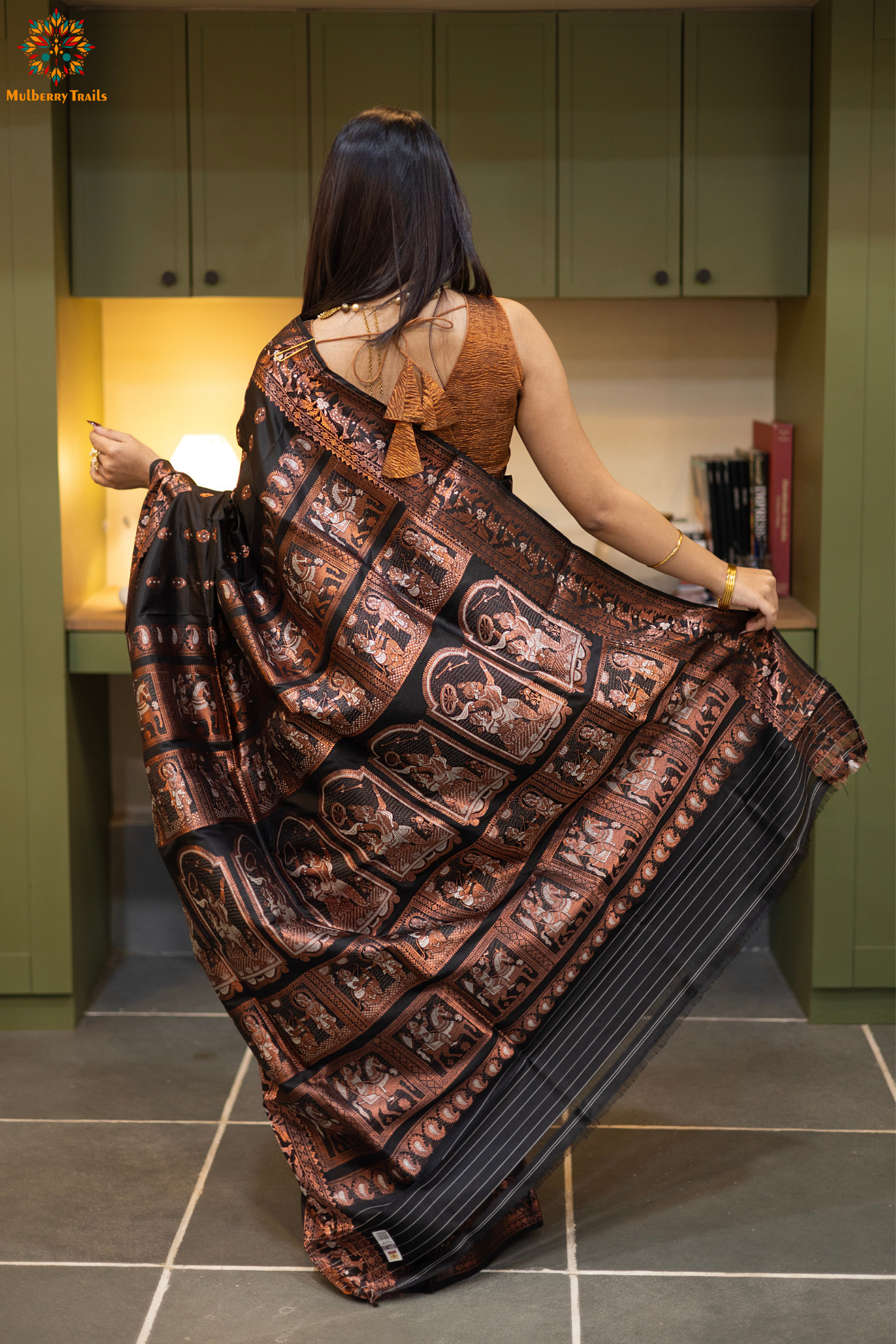 Swarnachari – Pure Silk Hazarbutta Saree
1. “Pure silk Swarnachari saree with hazarbutta thousand-motif weave and copper zari detailing for a regal look.”
2. “Silk Mark pure silk hazarbutta saree featuring intricate copper zari motifs and rich texture.”
Woman wearing a black and copper zari saree in a room with a desk and lamp.