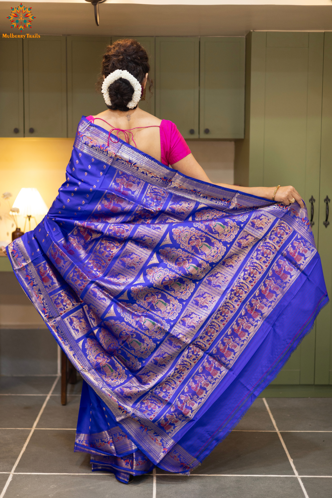 Swarnachari – Pure Silk Hazarbutta Saree
1. “Pure silk Swarnachari saree with hazarbutta thousand-motif weave and copper zari detailing for a regal look.”
2. “Silk Mark pure silk hazarbutta saree featuring intricate copper zari motifs and rich texture.”
Woman wearing a black and copper zari saree in a room with a desk and lamp.
