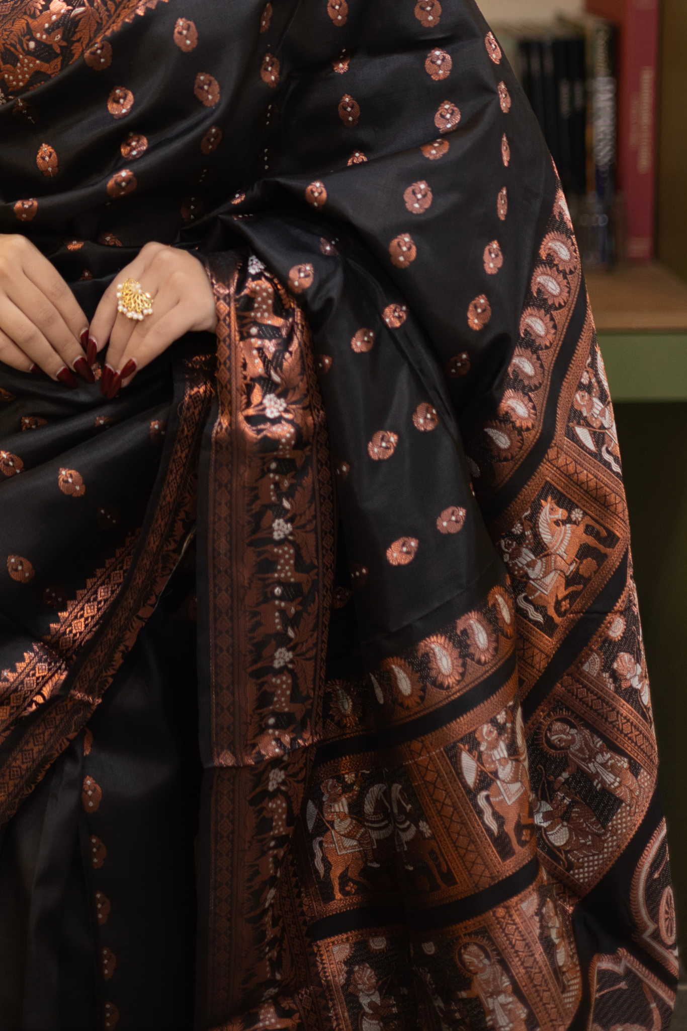Close-up of a black and brown patterned fabric with a hand wearing a ring.
Swarnachari – Pure Silk Hazarbutta Saree
1. “Pure silk Swarnachari saree with hazarbutta thousand-motif weave and copper zari detailing for a regal look.”
2. “Silk Mark pure silk hazarbutta saree featuring intricate copper zari motifs and rich texture.”
Woman wearing a black and copper zari saree in a room with a desk and lamp.
