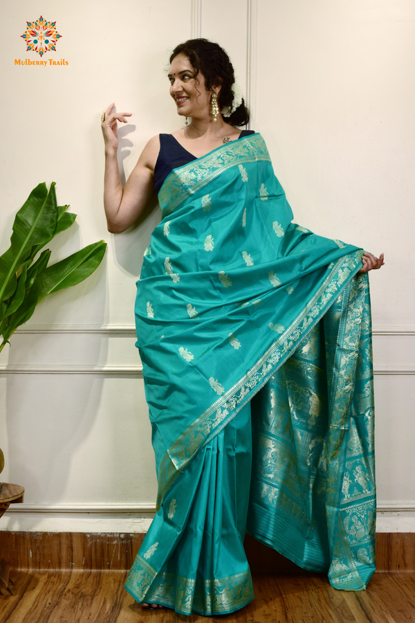 A woman wearing a Aqua Blue Baluchari Pure SIlk saree. purple baluchari silk , bengal silk saree with deer motif on pallu and ramayan, mahabharat motifs on pallu. This is a soft pure silk saree suitable for weddings, parties, festivities. suitable for Durga Puja shopping enthusiasts and diwali festival. 