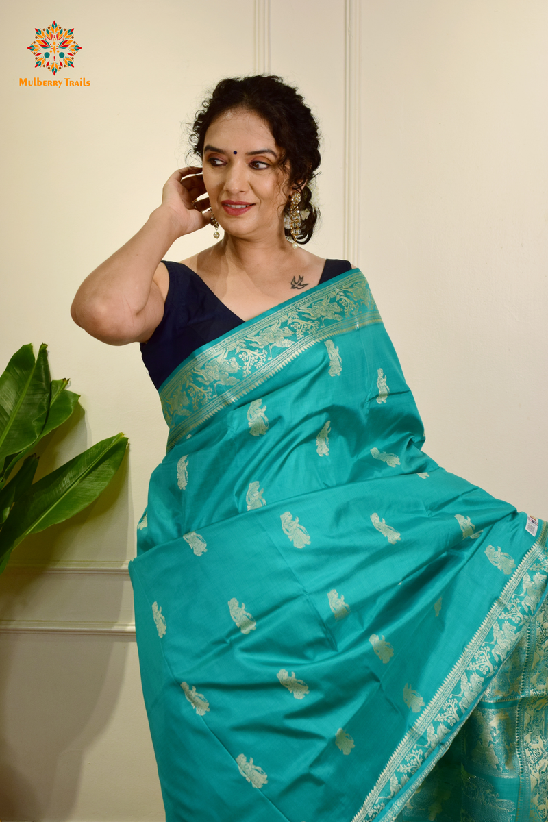 A woman wearing a Aqua Blue Baluchari Pure SIlk saree. purple baluchari silk , bengal silk saree with deer motif on pallu and ramayan, mahabharat motifs on pallu. This is a soft pure silk saree suitable for weddings, parties, festivities. suitable for Durga Puja shopping enthusiasts and diwali festival. 