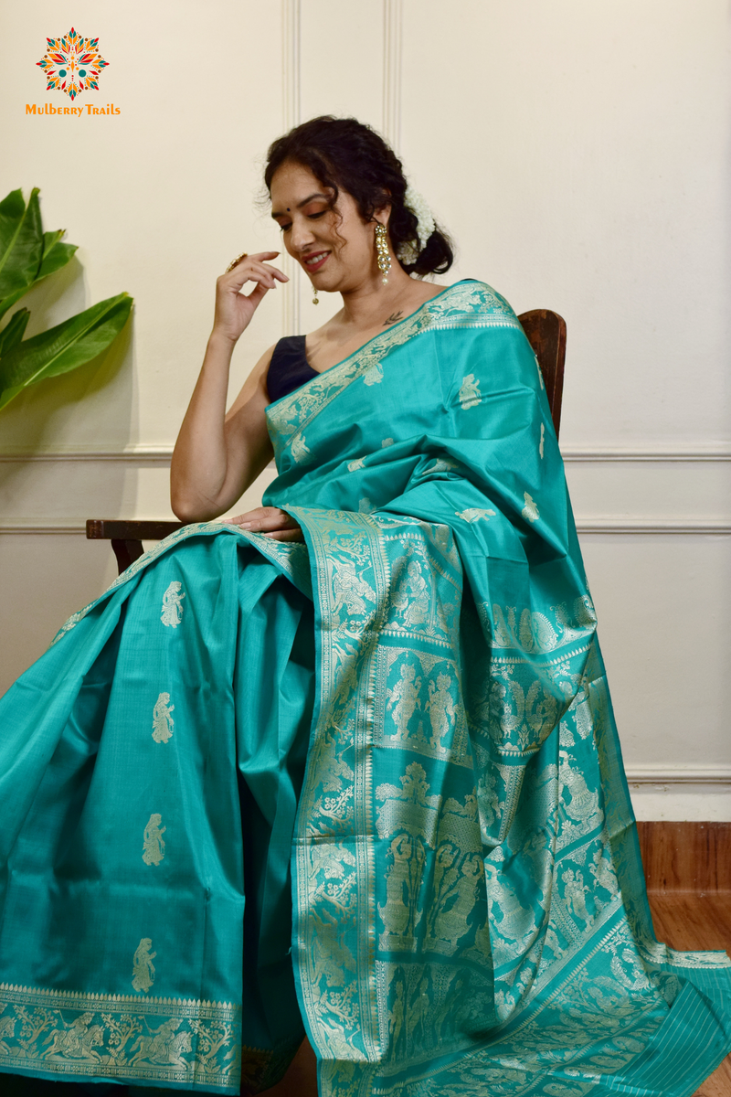 A woman wearing a Aqua Blue Baluchari Pure SIlk saree. purple baluchari silk , bengal silk saree with deer motif on pallu and ramayan, mahabharat motifs on pallu. This is a soft pure silk saree suitable for weddings, parties, festivities. suitable for Durga Puja shopping enthusiasts and diwali festival. 