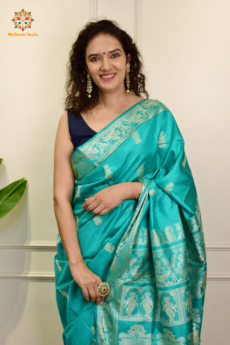 A woman wearing a Aqua Blue Baluchari Pure SIlk saree. purple baluchari silk , bengal silk saree with deer motif on pallu and ramayan, mahabharat motifs on pallu. This is a soft pure silk saree suitable for weddings, parties, festivities. suitable for Durga Puja shopping enthusiasts and diwali festival. 