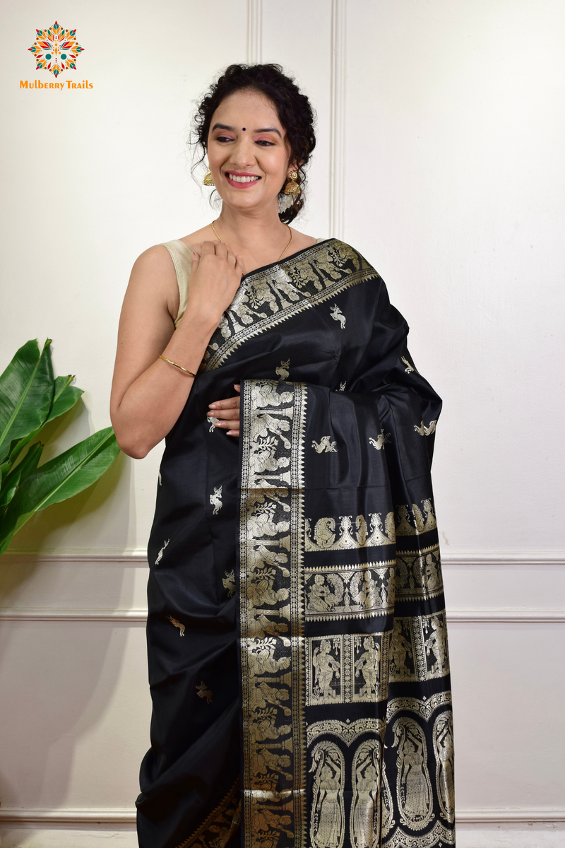 A woman wearing a black Baluchari Pure SIlk saree. Black baluchari silk , bengal silk saree with deer motif on pallu and ramayan, mahabharat motifs on pallu. This is a soft pure silk saree suitable for weddings, parties, festivities. suitable for Durga Puja shopping enthusiasts and diwali festival. 