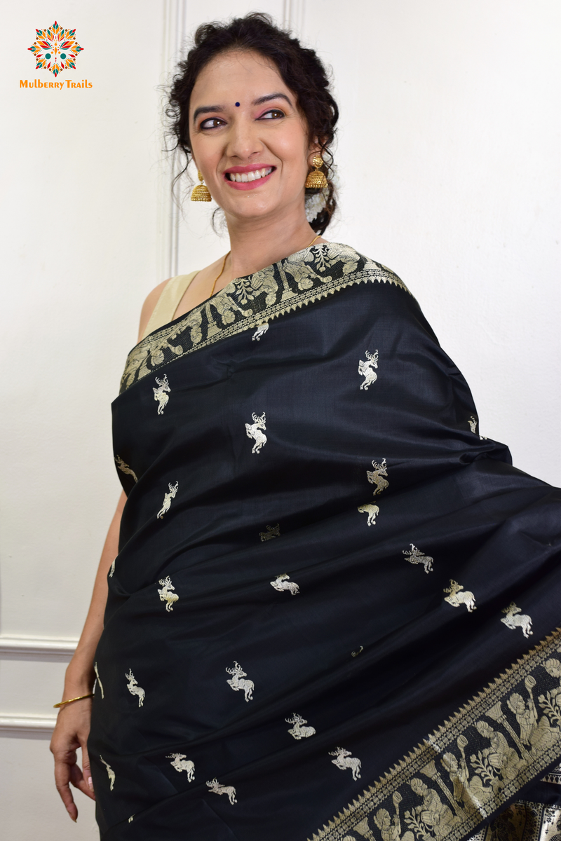 A woman wearing a black Baluchari Pure SIlk saree. Black baluchari silk , bengal silk saree with deer motif on pallu and ramayan, mahabharat motifs on pallu. This is a soft pure silk saree suitable for weddings, parties, festivities. suitable for Durga Puja shopping enthusiasts and diwali festival. 