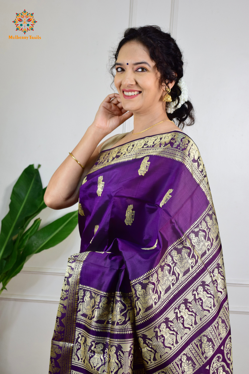 A woman wearing a Purple Baluchari Pure SIlk saree. purple baluchari silk , bengal silk saree with deer motif on pallu and ramayan, mahabharat motifs on pallu. This is a soft pure silk saree suitable for weddings, parties, festivities. suitable for Durga Puja shopping enthusiasts and diwali festival. 