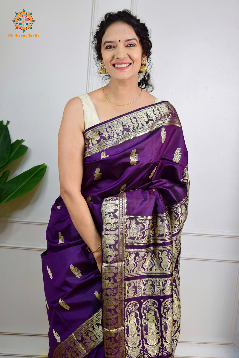 A woman wearing a Purple Baluchari Pure SIlk saree. purple baluchari silk , bengal silk saree with deer motif on pallu and ramayan, mahabharat motifs on pallu. This is a soft pure silk saree suitable for weddings, parties, festivities. suitable for Durga Puja shopping enthusiasts and diwali festival. 