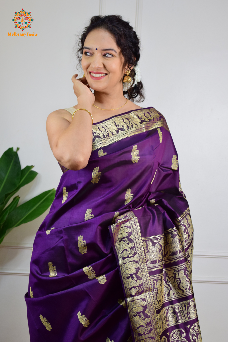 A woman wearing a Purple Baluchari Pure SIlk saree. purple baluchari silk , bengal silk saree with deer motif on pallu and ramayan, mahabharat motifs on pallu. This is a soft pure silk saree suitable for weddings, parties, festivities. suitable for Durga Puja shopping enthusiasts and diwali festival. 