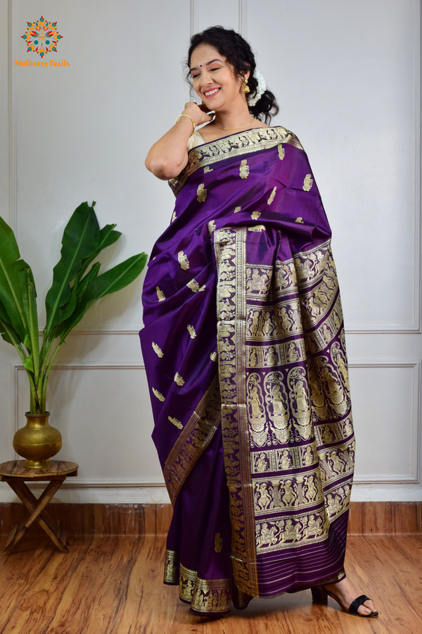 A woman wearing a Purple Baluchari Pure SIlk saree. purple baluchari silk , bengal silk saree with deer motif on pallu and ramayan, mahabharat motifs on pallu. This is a soft pure silk saree suitable for weddings, parties, festivities. suitable for Durga Puja shopping enthusiasts and diwali festival. 