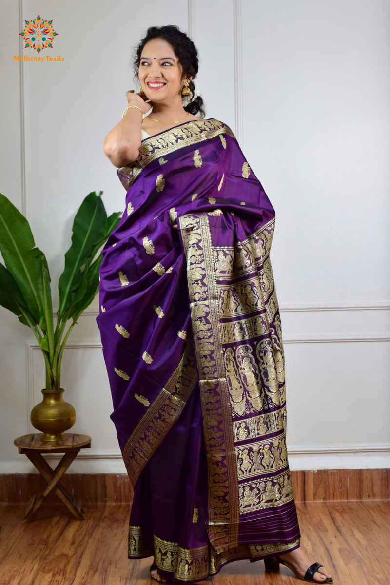 A woman wearing a Purple Baluchari Pure SIlk saree. purple baluchari silk , bengal silk saree with deer motif on pallu and ramayan, mahabharat motifs on pallu. This is a soft pure silk saree suitable for weddings, parties, festivities. suitable for Durga Puja shopping enthusiasts and diwali festival. 