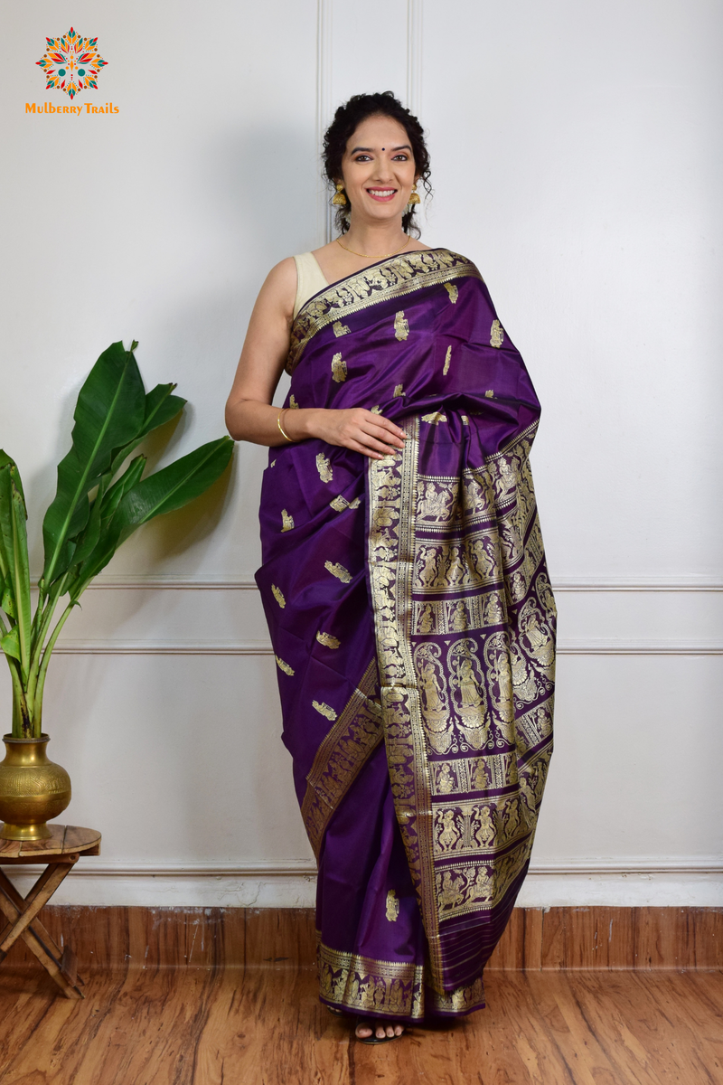 A woman wearing a Purple Baluchari Pure SIlk saree. purple baluchari silk , bengal silk saree with deer motif on pallu and ramayan, mahabharat motifs on pallu. This is a soft pure silk saree suitable for weddings, parties, festivities. suitable for Durga Puja shopping enthusiasts and diwali festival. 