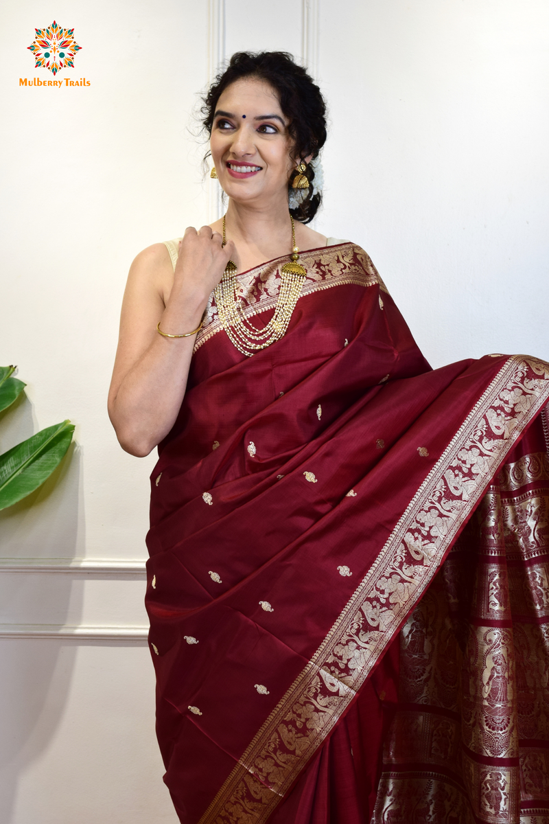 A woman wearing a Maroon Baluchari Pure SIlk saree. Maroon baluchari silk , bengal silk saree with deer motif on pallu and ramayan, mahabharat motifs on pallu. This is a soft pure silk saree suitable for weddings, parties, festivities. suitable for Durga Puja shopping enthusiasts and diwali festival. 