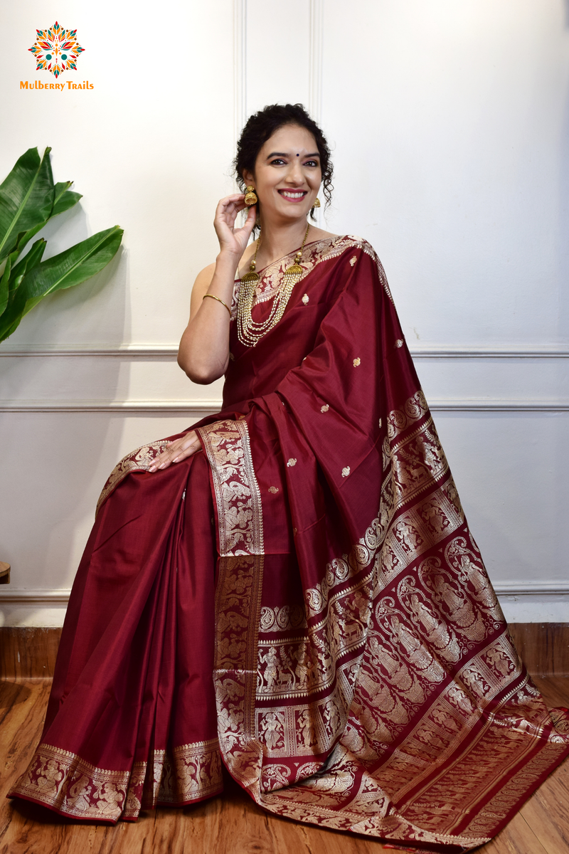 A woman wearing a Maroon Baluchari Pure SIlk saree. Maroon baluchari silk , bengal silk saree with deer motif on pallu and ramayan, mahabharat motifs on pallu. This is a soft pure silk saree suitable for weddings, parties, festivities. suitable for Durga Puja shopping enthusiasts and diwali festival. 