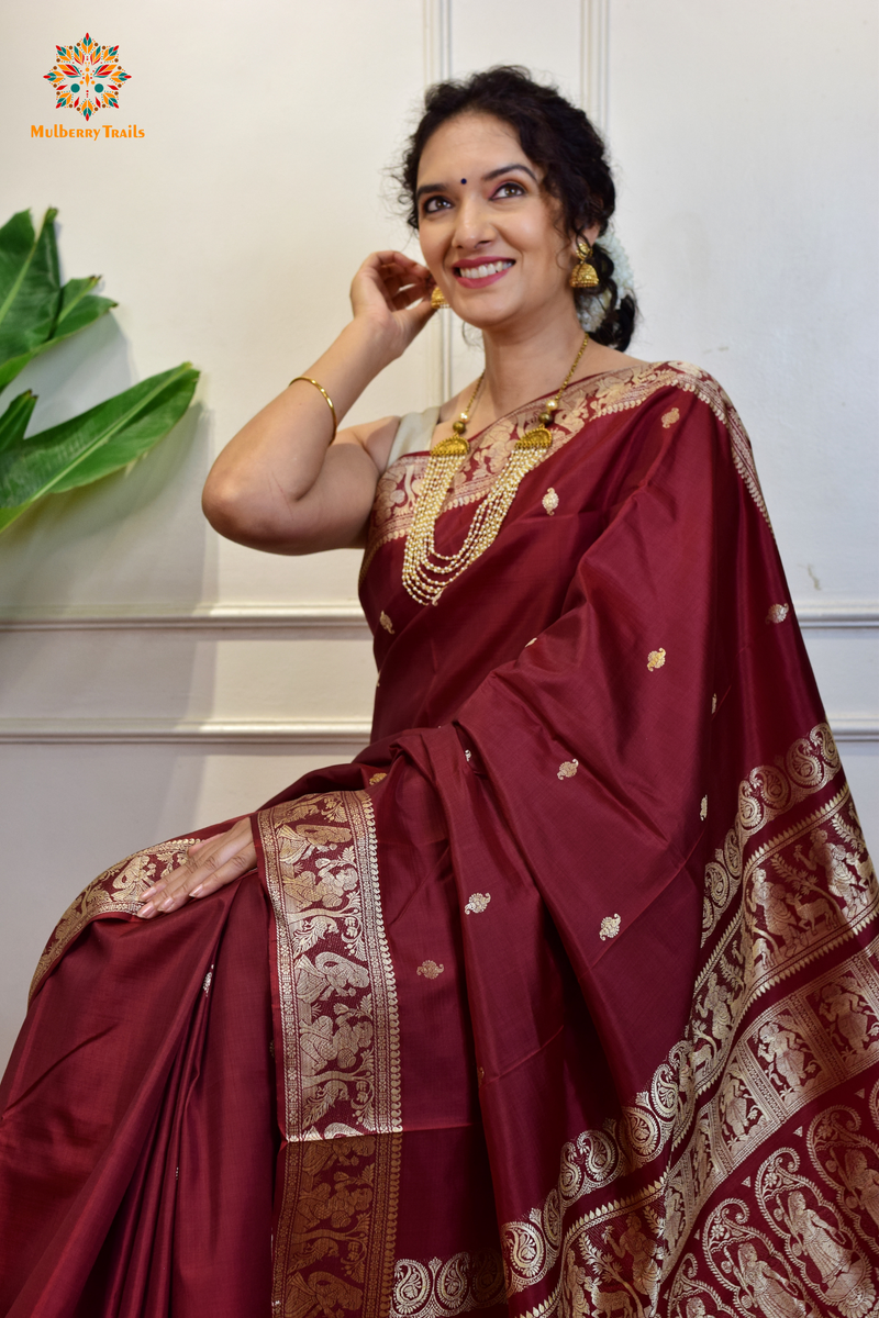 A woman wearing a Maroon Baluchari Pure SIlk saree. Maroon baluchari silk , bengal silk saree with deer motif on pallu and ramayan, mahabharat motifs on pallu. This is a soft pure silk saree suitable for weddings, parties, festivities. suitable for Durga Puja shopping enthusiasts and diwali festival. 