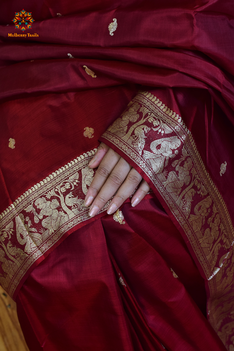 A woman wearing a Maroon Baluchari Pure SIlk saree. Maroon baluchari silk , bengal silk saree with deer motif on pallu and ramayan, mahabharat motifs on pallu. This is a soft pure silk saree suitable for weddings, parties, festivities. suitable for Durga Puja shopping enthusiasts and diwali festival. 